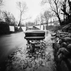 A pinhole captures a black and white photo of a piano gracefully placed on the side of a serene river